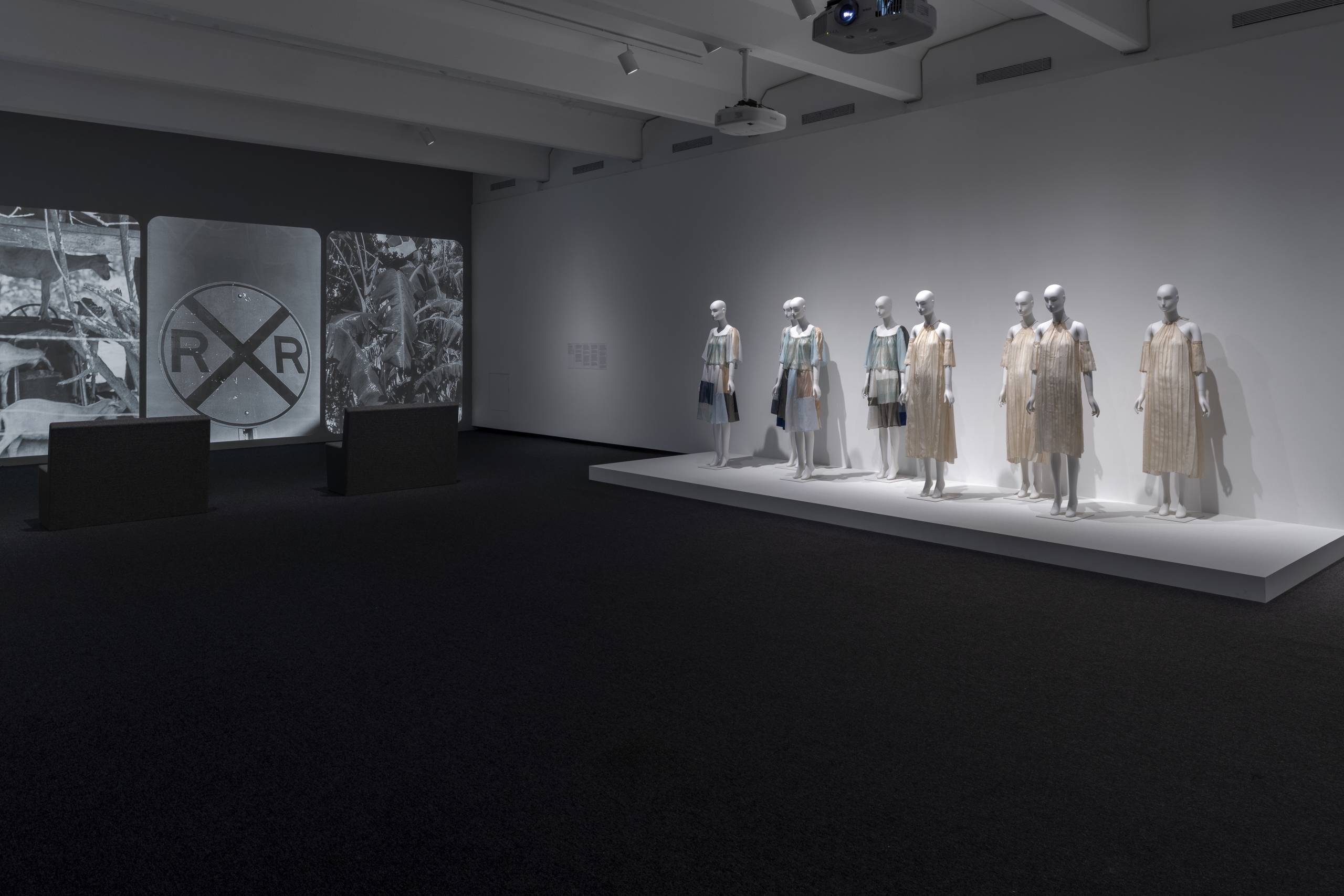 Exhibition gallery with six mannequins wearing dresses on a platform, an image of a vintage RXR railroad crossing projected on the wall, and two rectangular dark benches in the foreground.
