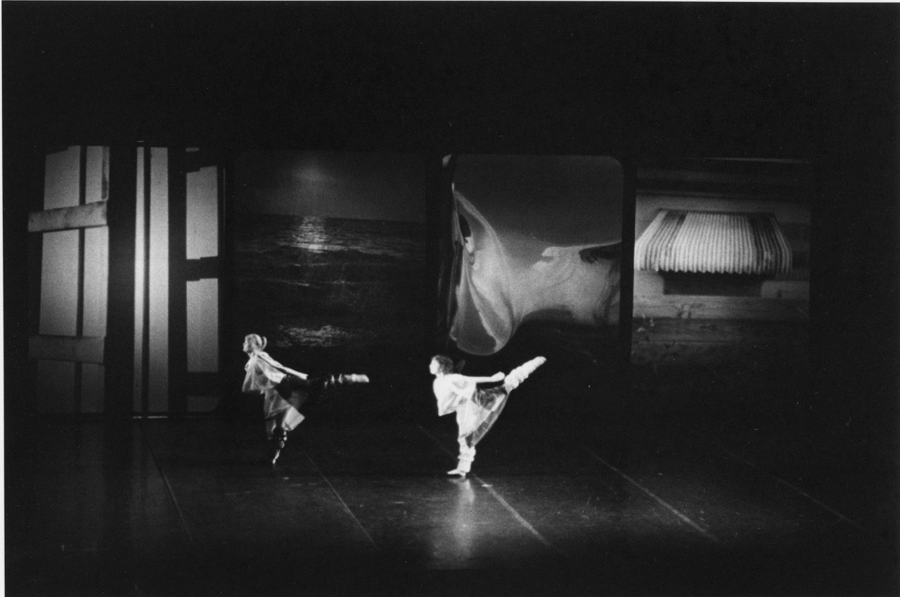 Two dancers on a stage hold matching poses, each standing on one leg with the other stretched straight out. Behind them, large rectangular screens display different images, including ocean waves, flowing fabric, and close-up portions of buildings. The lighting highlights the dancers while the rest of the stage remains dim.