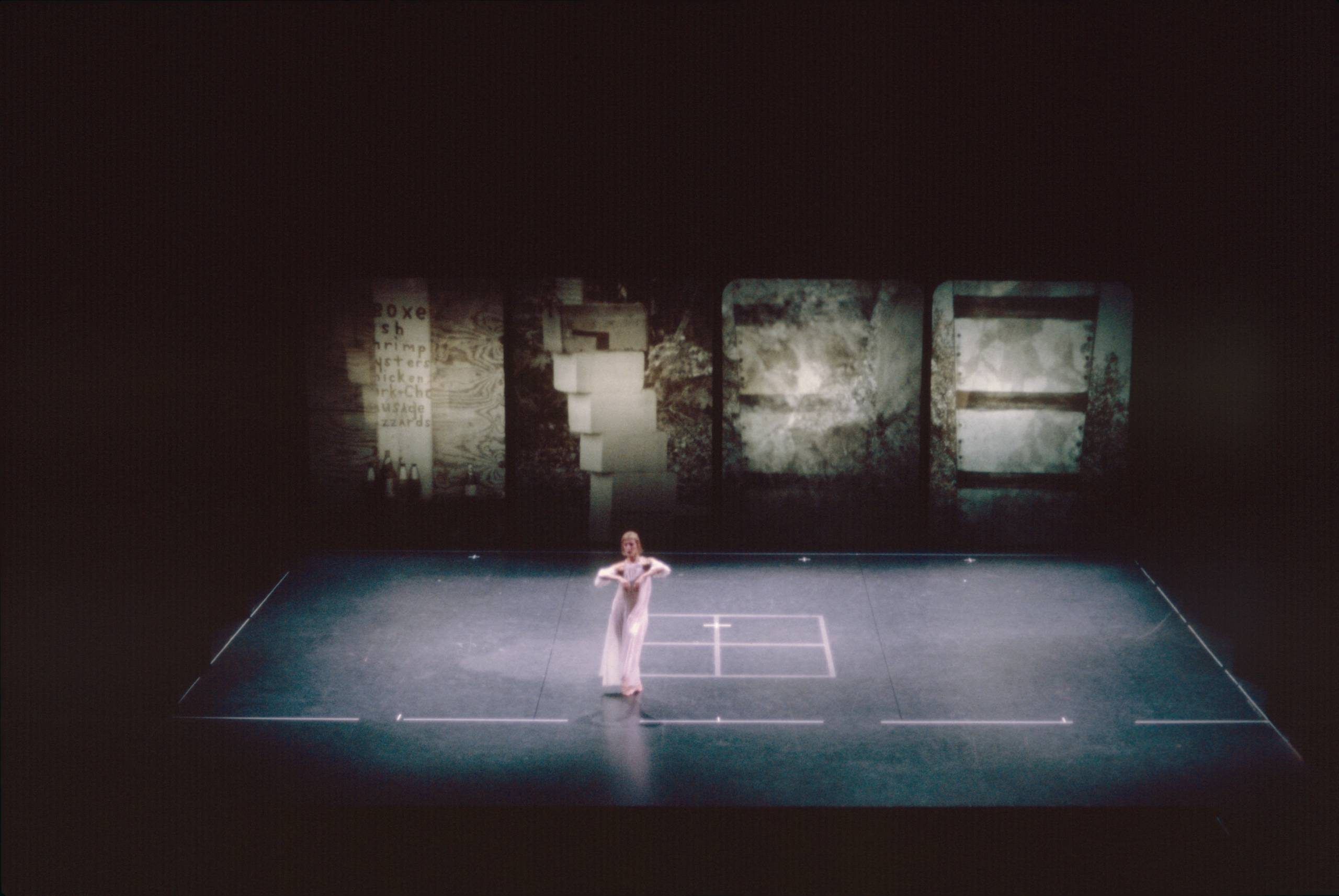 A dancer stands alone on a dimly lit stage, centered in front of a large projection of four close-up, abstract details of structures. The stage is marked with white lines.