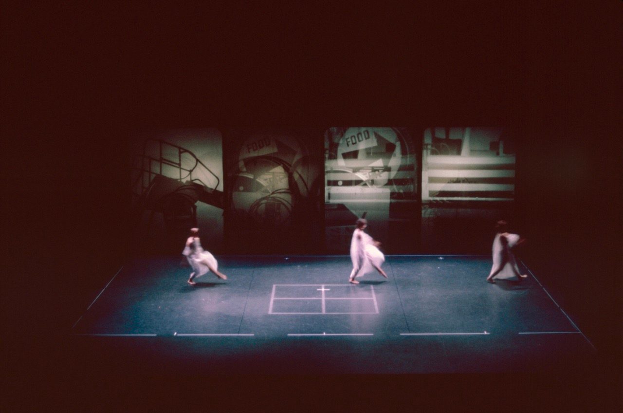 Wide view of a theater stage showing three performers in light-colored costumes moving in different directions across a dark floor. A square marking is centered on the stage, with large illuminated panels and abstract imagery in the background.