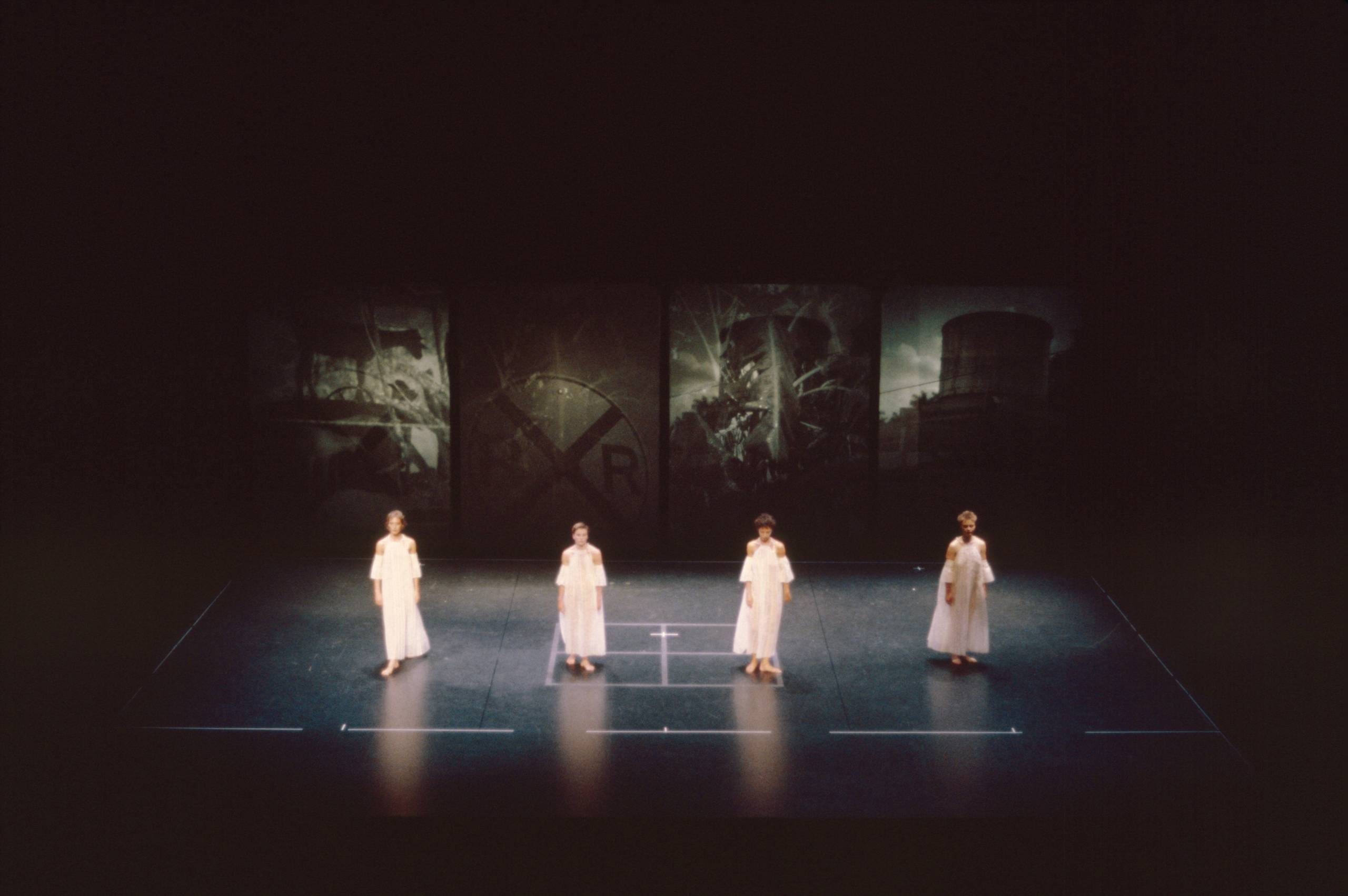 Four people in white outfits stand spaced apart on a dark stage with faded industrial images in the background. The stage is marked with white lines, and the lighting highlights the performers.