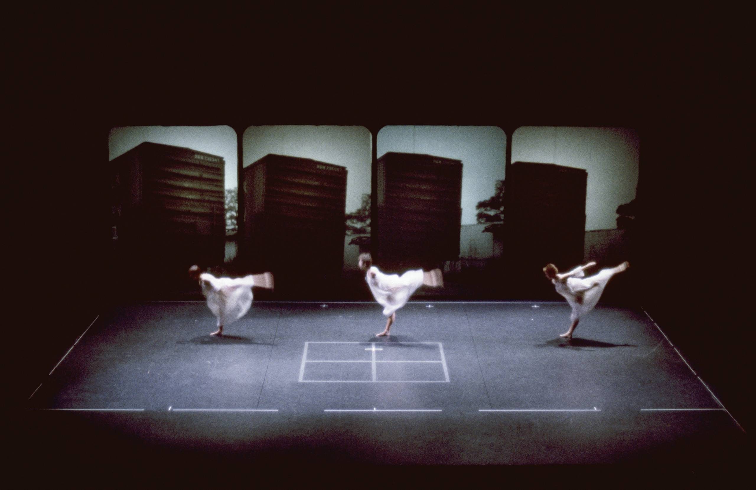 Three dancers in white costumes perform synchronized movements on a dark stage, each standing on one leg with arms extended. Large screens in the background display images of tall structures. The stage has white lines and a square.