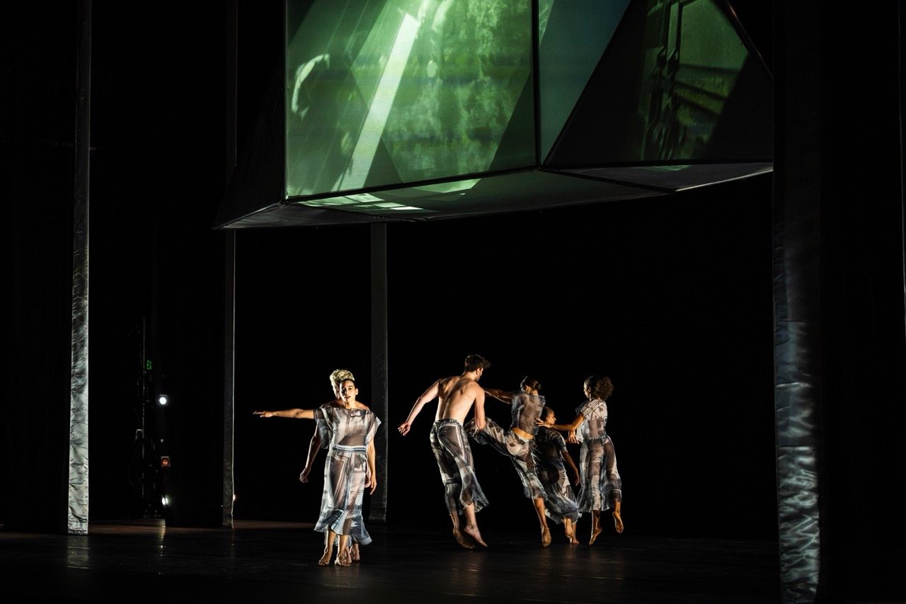 Five dancers in translucent, flowing costumes move across a dimly lit stage beneath a large hanging installation with projected green-tinted video imagery. Tall vertical set elements frame the space.