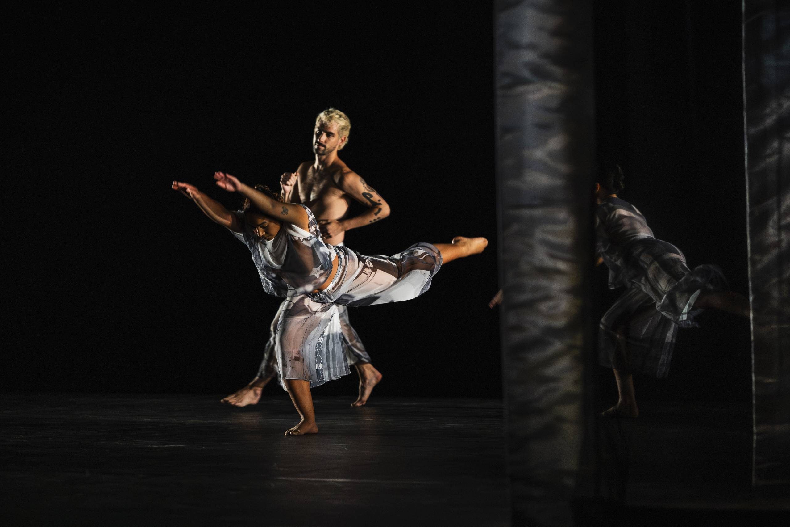 Two dancers perform on a dimly lit stage; one balances on one leg with arms outstretched, while the other stands behind, both wearing translucent costumes. Reflections are visible on a nearby mirrored surface.