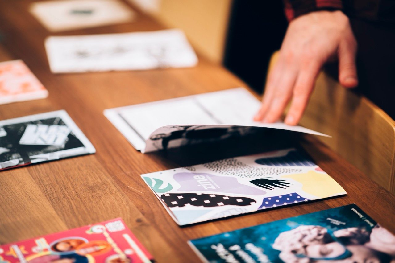 A person browses colorful magazines and zines laid out on a wooden table, with one hand flipping through the pages of a booklet.