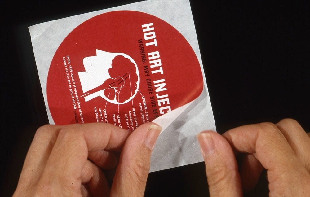 A person peels a red-and-white sticker labeled HOT ART INJECTION, WARNING: MAY CAUSE SIDE EFFECTS, with a graphic of a head labeled with additional side effect information.