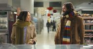 A woman and a bearded man wearing winter coats and scarves smile at each other while standing in a grocery store aisle decorated with holiday ornaments.