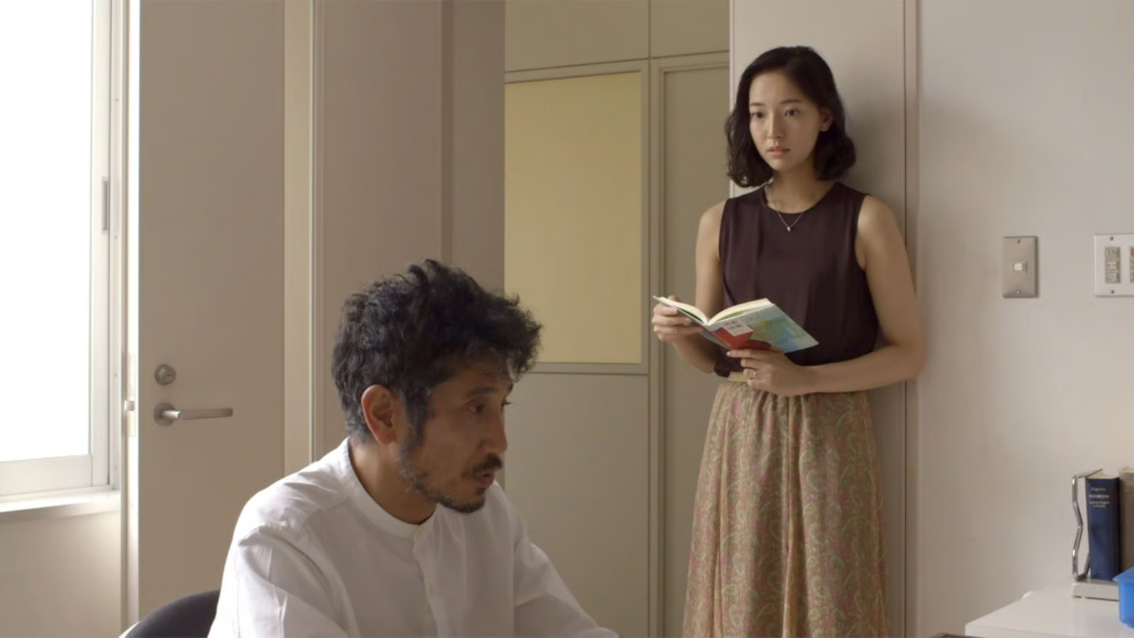 A man sits at a desk looking serious while a woman stands behind him by the door, holding an open book and watching him quietly.