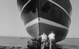 Two people work on the bow of a large boat that is docked on land near the shore. The photo is black and white, and the boat dominates the foreground with the sea and horizon in the background.