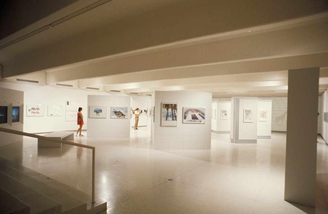 A spacious, modern art gallery with white walls, various artworks displayed, and a few visitors, including a woman in a red dress.