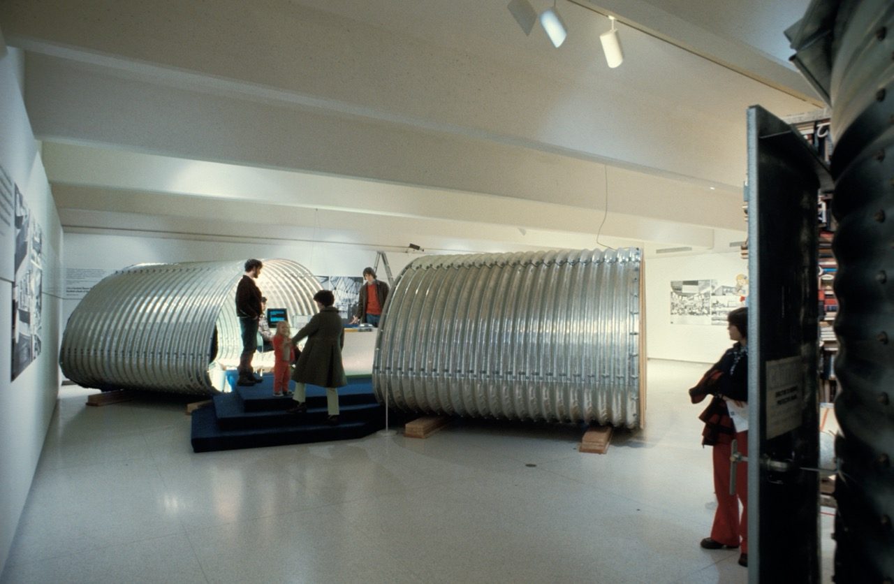 A group of people explores a museum exhibition featuring two large, ribbed metal capsule structures displayed in a spacious, well-lit gallery.