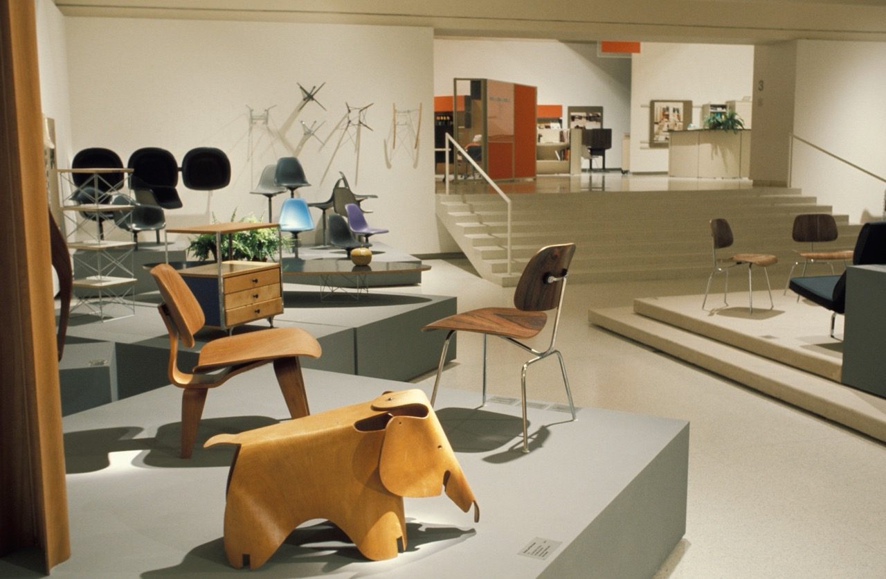 Museum exhibition featuring mid-century modern furniture, including wooden chairs, a small drawer unit, and a wooden elephant-shaped stool, all displayed on platforms in a spacious, well-lit gallery.