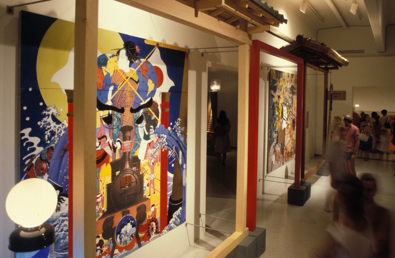Colorful Japanese artwork displayed in a museum exhibition, featuring bold figures and vibrant patterns. Visitors walk through the gallery, observing the art, which is framed by traditional architectural elements.