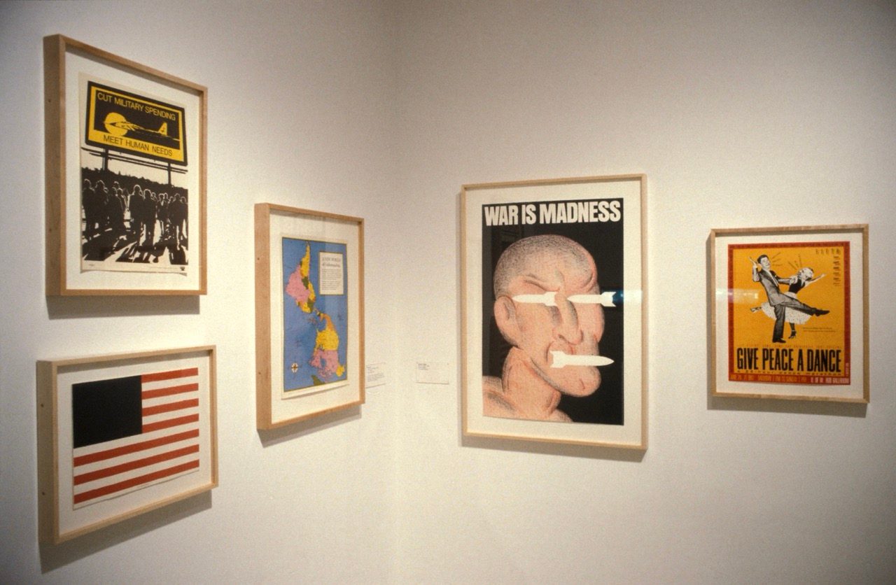 Five framed posters with anti-war and peace messages hang on a white wall, including images of a flag, protestors, a map, a dancing couple, and a stylized face with the caption 