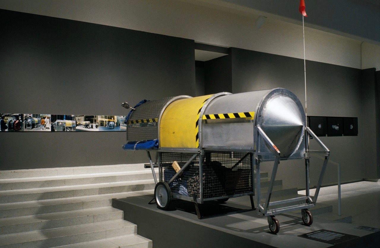 A large, cylindrical, industrial-looking mobile shelter with wheels is displayed in a gallery on a platform, near a series of small photographs on the wall.