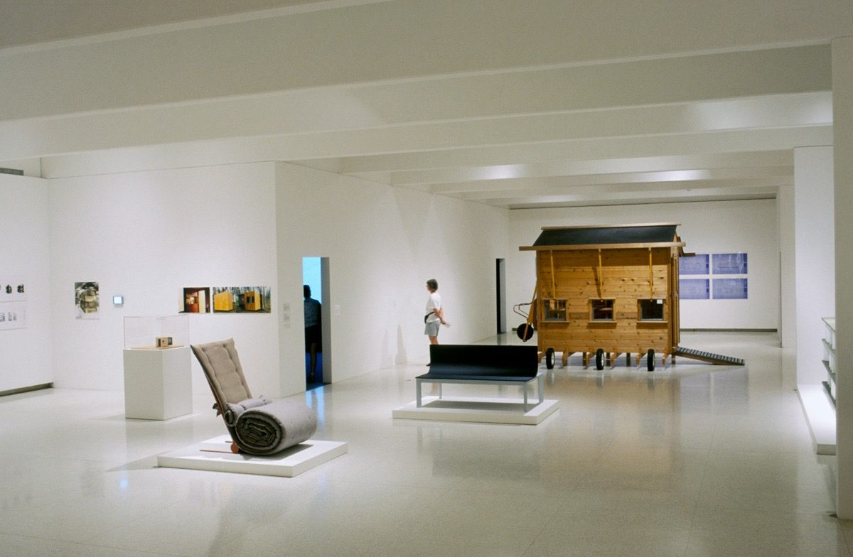 A modern art gallery with white walls, displaying unique pieces of furniture and a large wooden structure; a person stands near an open doorway in the background.