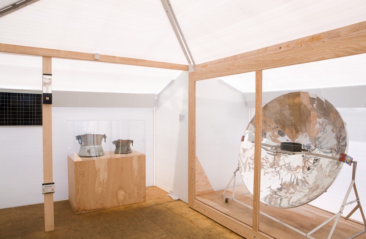 A bright room with wooden framing featuring two metal pots displayed on a wooden pedestal and a large reflective concave dish on a stand.