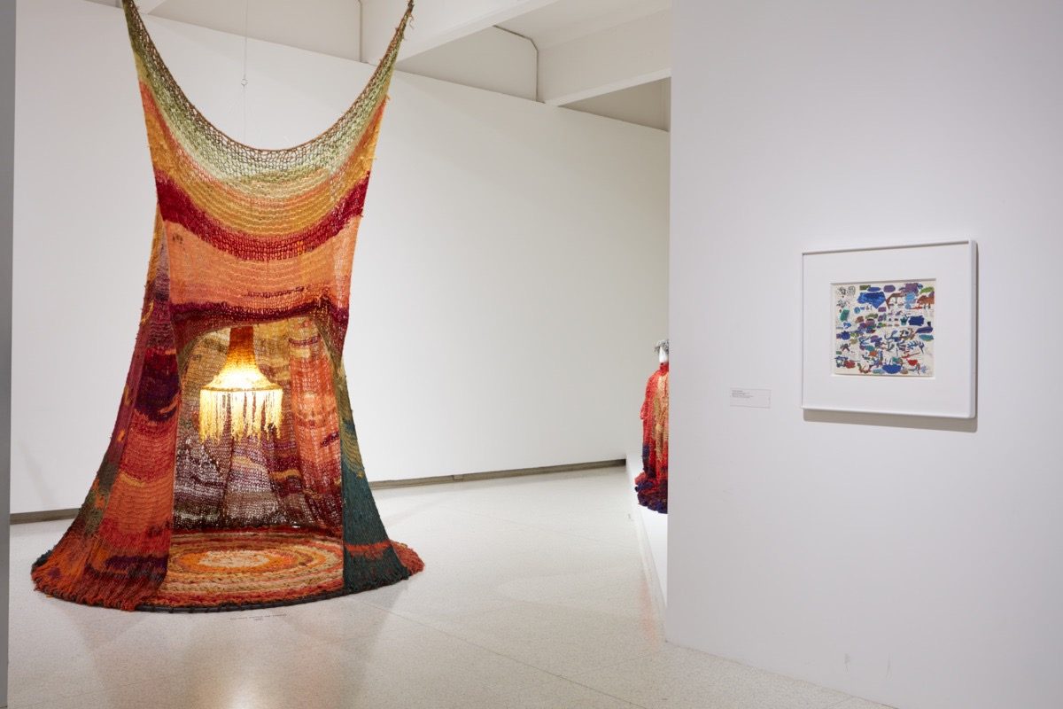 A colorful, tent-like fiber art installation with warm lighting inside installed in a gallery space. On the right wall is a small framed artwork featuring abstract patterns.