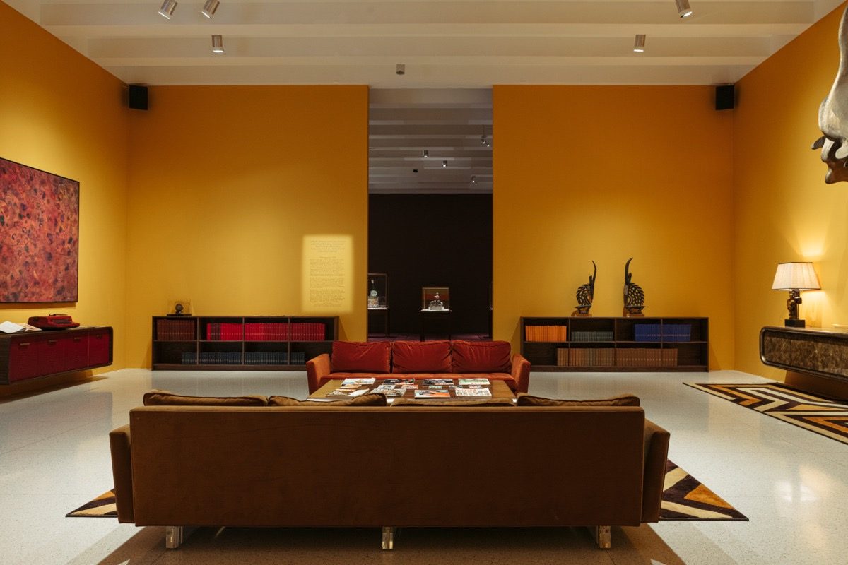 A gallery installed as a modern living room with mustard yellow walls, brown sofas, a coffee table with magazines, wall art, shelves, decorative sculptures, and soft lighting. The space feels warm and inviting.