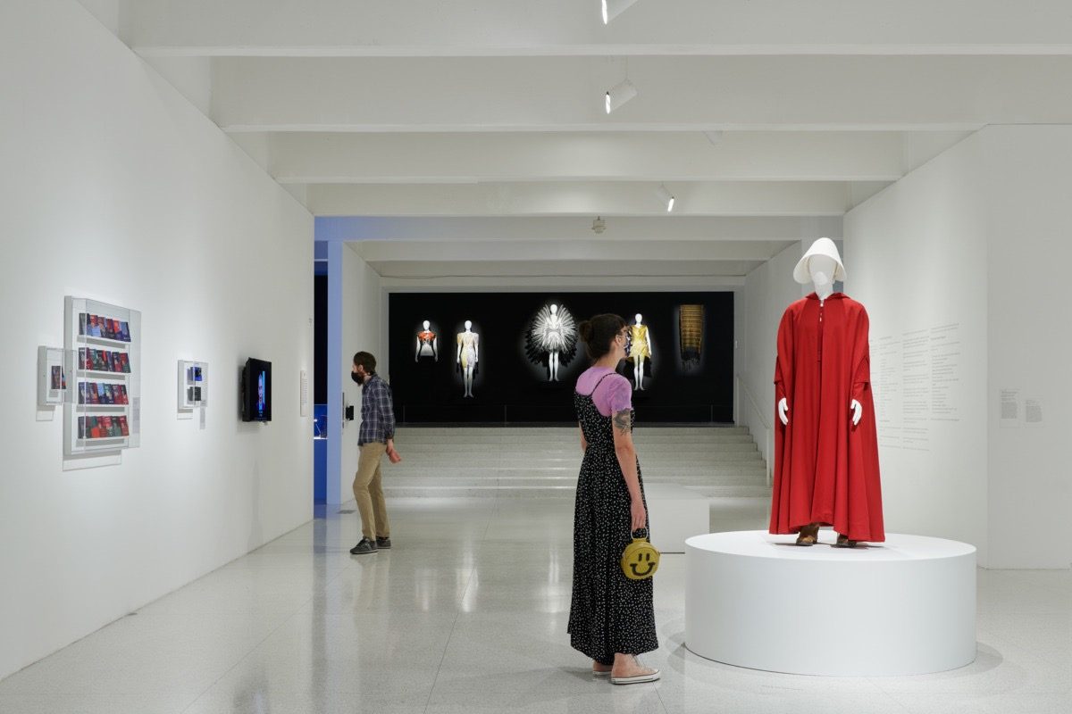A woman looks at a red cloak and white bonnet displayed on a mannequin in a modern art gallery, with another visitor views a video displayed alongside various artworks on the walls.