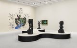 A modern art gallery with abstract sculptures on a black and white display, a wall mural with green, black, and blue designs, and a framed digital artwork on the wall. The room has bright lighting and a white floor.