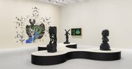 A modern art gallery with abstract sculptures on a black and white display, a wall mural with green, black, and blue designs, and a framed digital artwork on the wall. The room has bright lighting and a white floor.