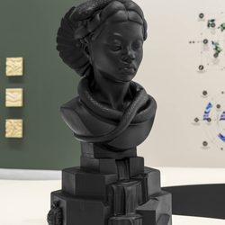 A matte black bust of a woman with intricate hair, a snake wrapped around her neck, and geometric shapes at the base. The background is a gallery wall with some small square and abstract artworks.