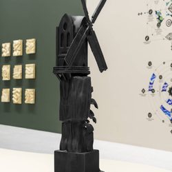 A black sculpture of a windmill atop a stylized, stacked, hand-like base is displayed indoors, with abstract art pieces and diagrams visible on the walls behind it.