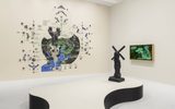 Gallery with a wall mural featuring diagrams and symbols, a black abstract windmill-like sculpture on a curved platform, and a framed digital artwork with green, black, and white graphics hanging on the side wall.