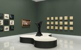 An art gallery room with dark green walls displays framed artworks, a central black abstract sculpture on a white platform, and a grid of textured beige panels on the right wall.