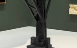 A black sculptural artwork resembling a broken or unfinished elevated railway stands on a rocky base in a gallery, with art pieces visible on the green walls behind it.