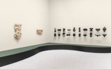 A minimalist gallery features beige walls with two tan abstract sculptures and a grid of small, black, organic-shaped pieces. A curved, green and black bench runs along the walls.