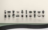 Twenty black sculptural masks hang in a horizontal row on a white wall, casting distinct shadows below each piece. The floor has curved sections with green and black coloring.