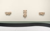 Three abstract wooden sculptures with organic, carved patterns are mounted on a light beige wall above a green wavy stripe and white tiled floor in a minimalist gallery setting.