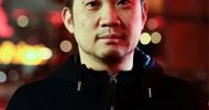 Headshot of Ryusuke Hamaguchi outdoors in a city at night. There are out of focus lights in red and yellow in the background.