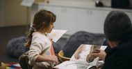 A young girl holds a sheet of paper while an adult points to a page in an open picture book. They are sitting at a table in a cozy indoor setting with books and cushions around.