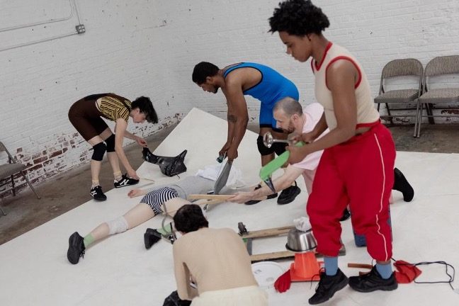 A group of people in a white studio space interact with various objects and a person lying on the floor.