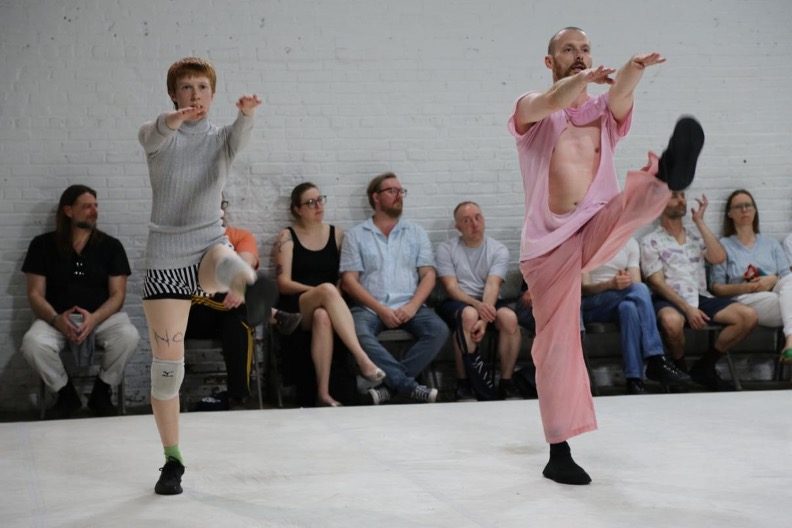 Two dancers perform a movement with extended legs in front of a seated audience.