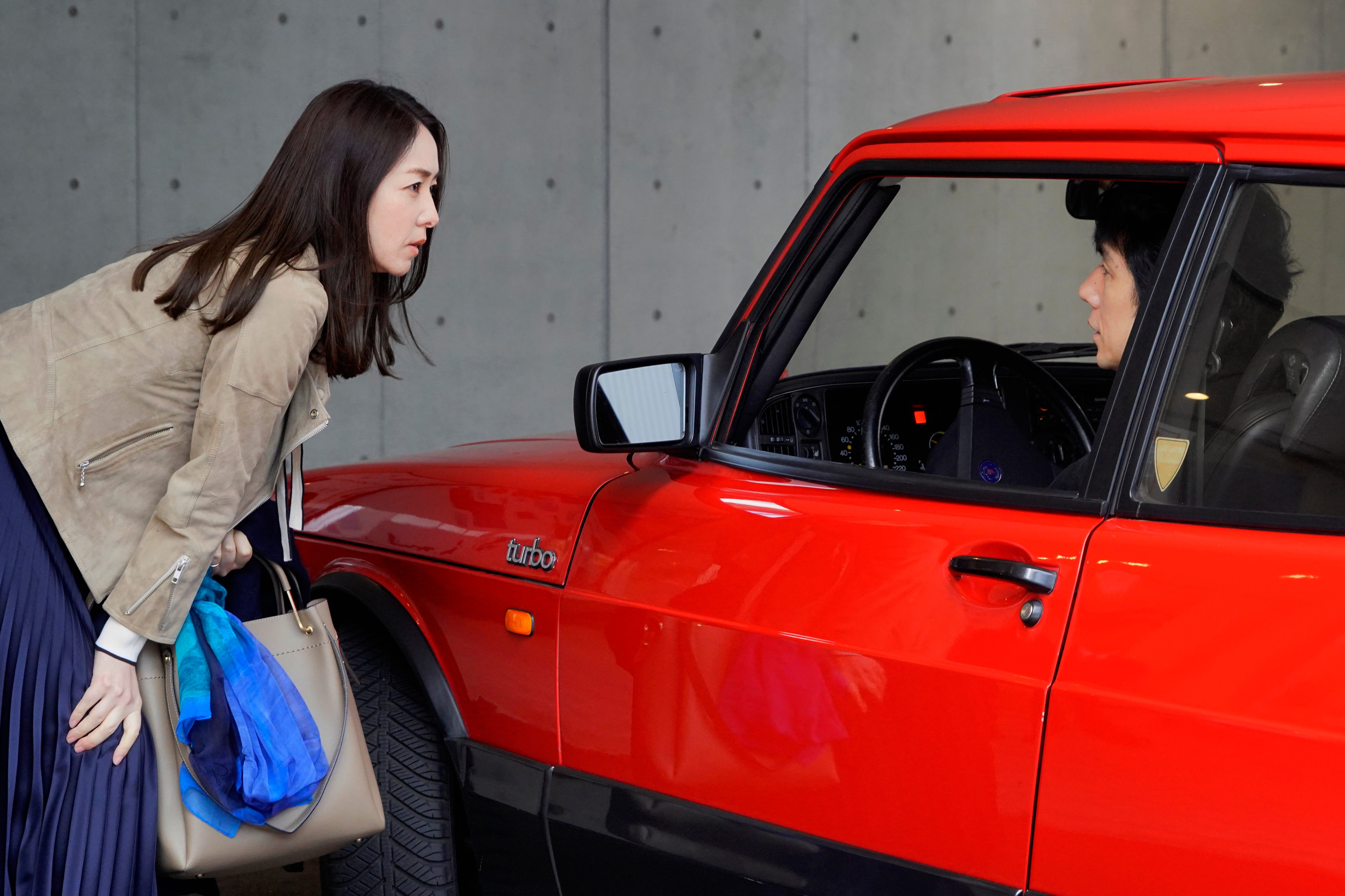 A person carrying a handbag and blue scarf leans toward the open window of a bright red car, speaking with the driver inside. In the background is a plain gray concrete wall.
