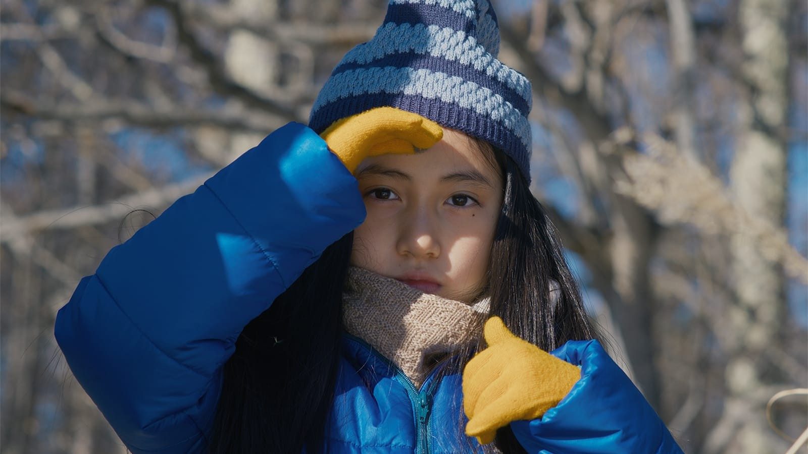 A young girl wearing a blue winter coat, a striped beanie, and yellow mittens shields her eyes from the sun.