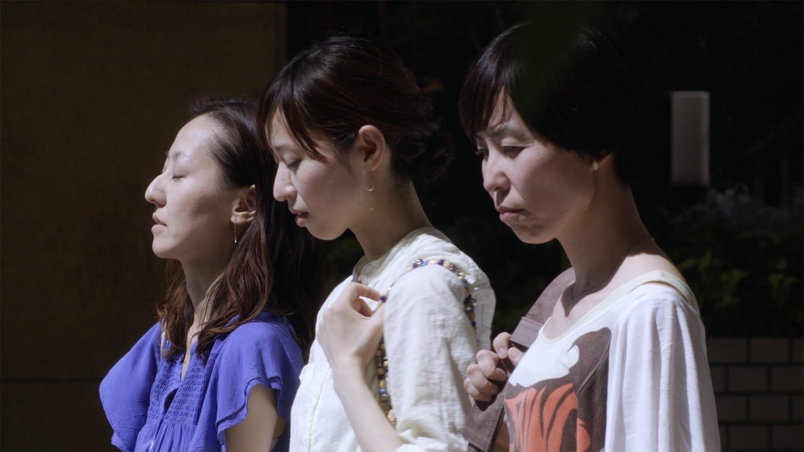 Three women, appearing in profile from the chest up, walk in a line again a dark background.